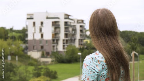 A beautiful young Caucasian woman looks at an apartment complex in a suburban area - rear view