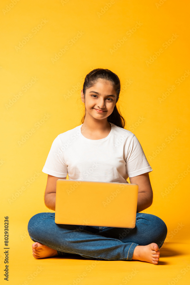 portrait of Indian asian schoolgirl girl using laptop, sitting cross-legged isolated on yellow background portrait of Indian asian schoolgirl girl using laptop, sitting cross-legged isolated on yellow background