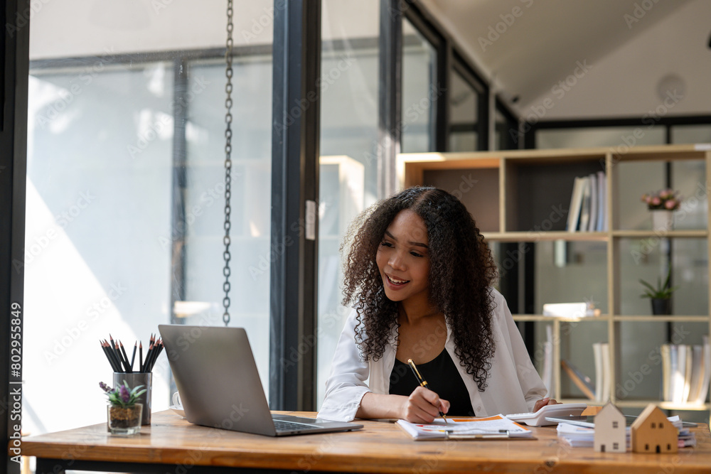 A woman is sitting at a desk with a laptop and a pen. She is smiling and she is working on something. The scene suggests a positive and productive atmosphere A woman is sitting at a desk with a laptop and a pen. She is smiling and she is working on something. The scene suggests a positive and productive atmosphere
