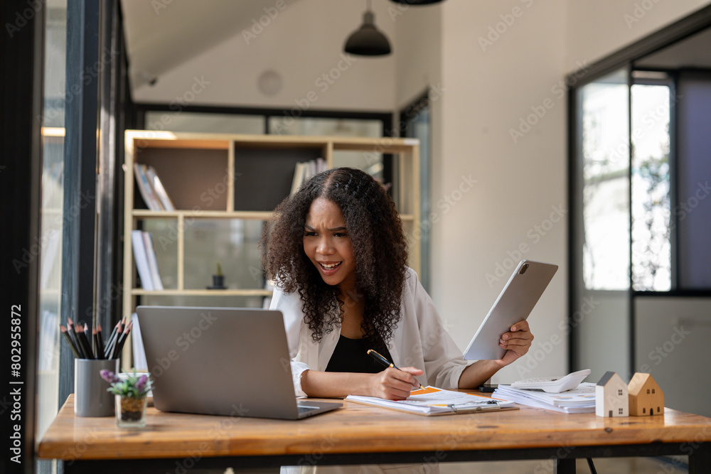 © Wasana - A woman is sitting at a desk with a laptop and a tablet. She is holding a tablet in one hand and a pen in the other. She is frustrated or angry, possibly due to a work-related issue © Wasana - A woman is sitting at a desk with a laptop and a tablet. She is holding a tablet in one hand and a pen in the other. She is frustrated or angry, possibly due to a work-related issue