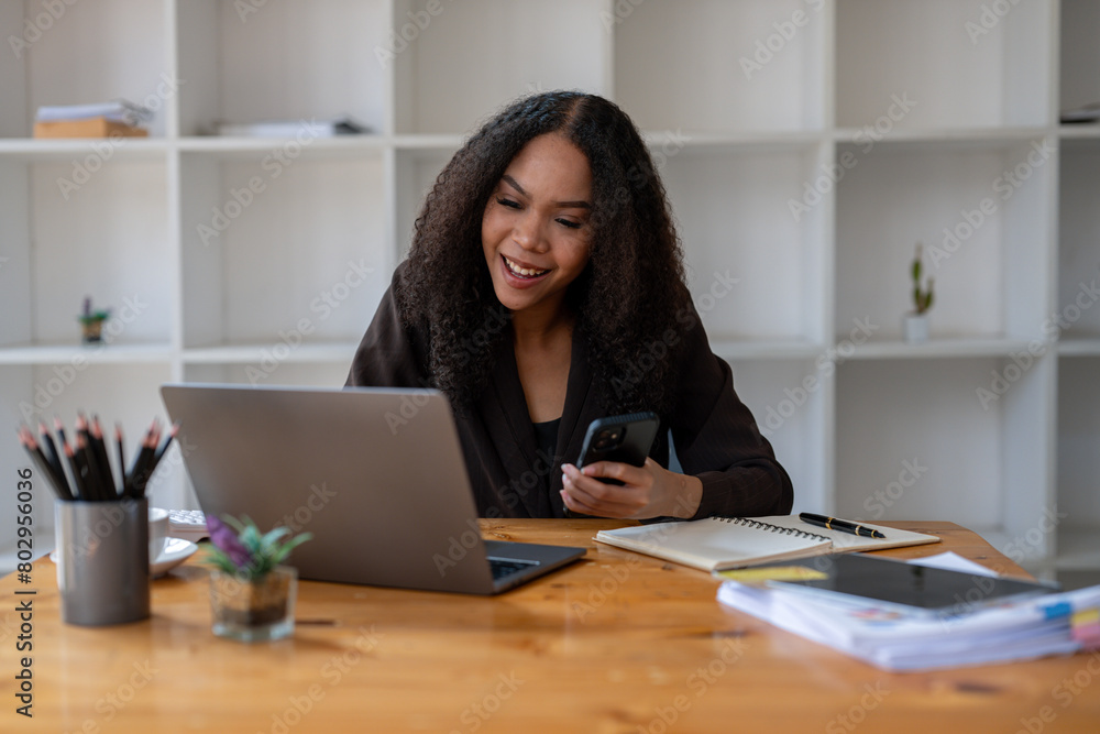 A woman is sitting at a desk with a laptop and a cell phone. She is smiling and she is happy A woman is sitting at a desk with a laptop and a cell phone. She is smiling and she is happy