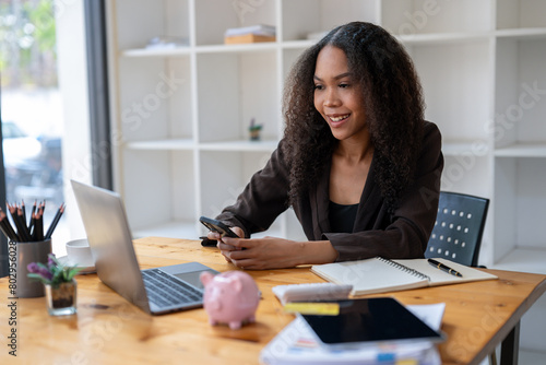 A woman is sitting at a desk with a laptop and a pink piggy bank. She is smiling and holding a cell phone. The scene suggests a relaxed and comfortable atmosphere © Wasana