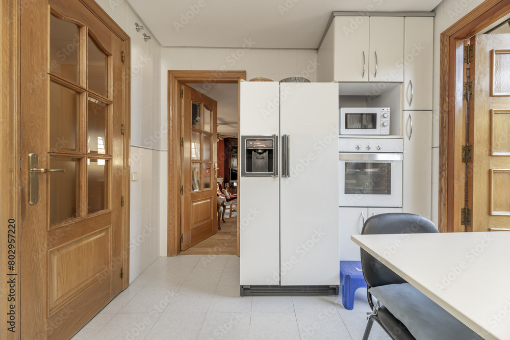 Frontal image of a kitchen with white furniture with metal handles and a column with a refrigerator and conventional and microwave ovens Frontal image of a kitchen with white furniture with metal handles and a column with a refrigerator and conventional and microwave ovens