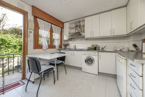 Frontal image of a kitchen with white furniture with metal handles, gray countertops and wooden windows and access to an outside plot