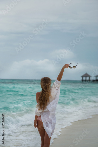 Beautiful girl with long blond hair. A girl stands by the ocean and looks into the distance. The girl is holding sunglasses in her hands. The wind blows my hair and white beach clothes. Beautiful tan.