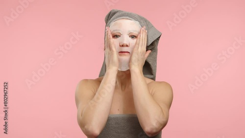 A young Caucasian woman wearing a gray towel headwrap applying a moisturizing sheet mask on her face against a soft pink background. Perfect for beauty and skincare promotions. 