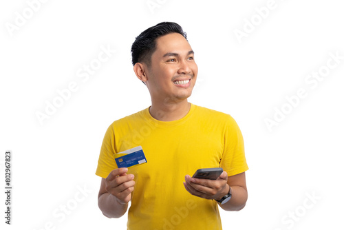 Happy young Asian man holding mobile phone and bank credit card, looking aside with big smile isolated on white background © Sewupari Studio