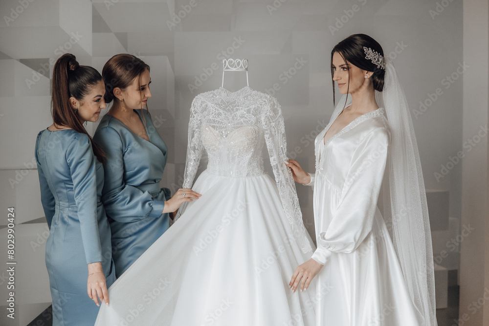 Three women are standing in front of a white wedding dress. One of the women is holding the dress up to the other two. Scene is one of excitement and anticipation for the upcoming wedding