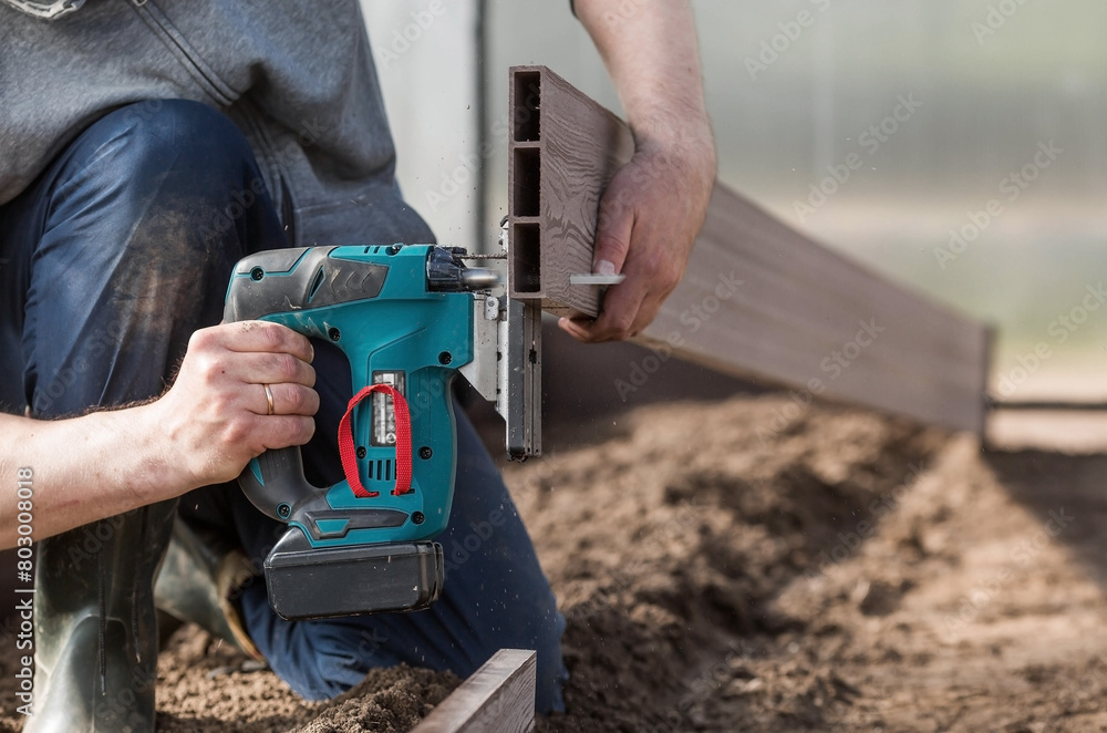 A man holds a battery-powered electric saw and builds new beds in a greenhouse made of wood-polymer composite. Macro photography. Side view. Preparing for the spring garden season. A man holds a battery-powered electric saw and builds new beds in a greenhouse made of wood-polymer composite. Macro photography. Side view. Preparing for the spring garden season.