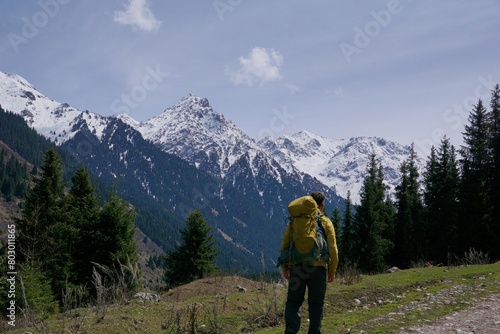 a man with a tourist backpack walks along a mountain road against the background of a forest and snowy mountains