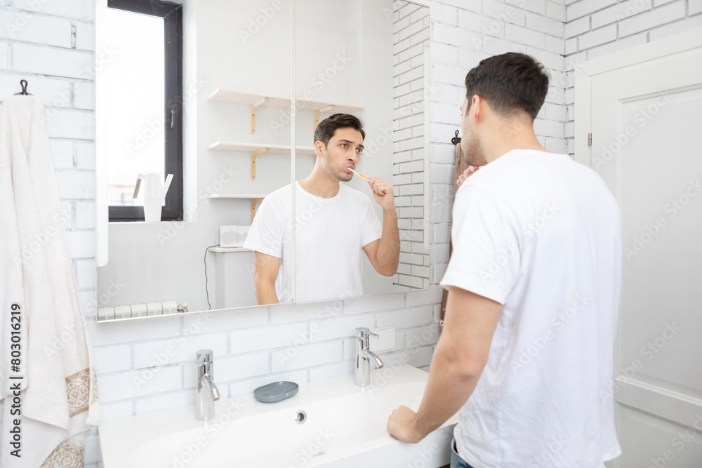 Fototapeta premium Handsome man brushing teeth and looking at the mirror in the bathroom 