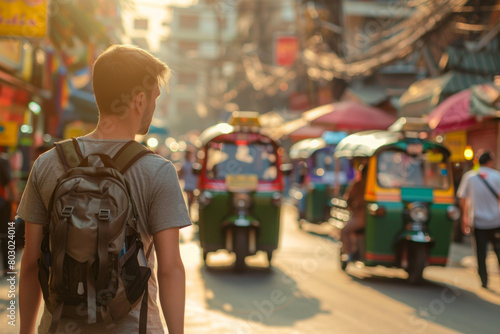 Man in t-shirt with a backpack from his back walks along asian street. Back view of a man. Tuk tuks and an asian market on a blurred background. Daytime. Copy space photo