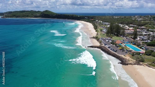 Drone flying along coast with view of surrounding buildings, waves, beach and mountains in distance photo