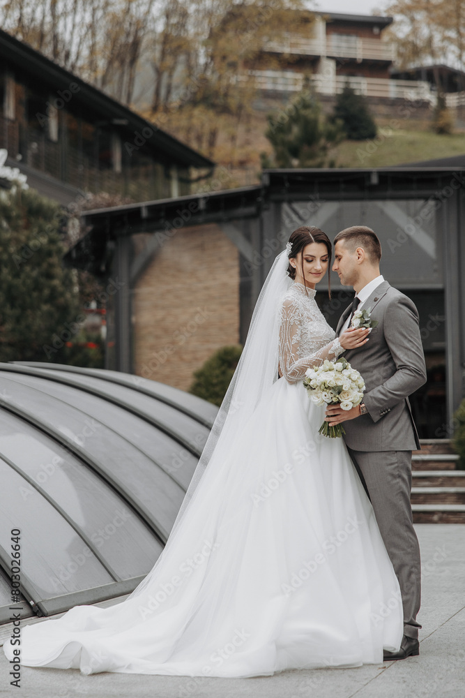 A bride and groom are posing for a picture in front of a building. The bride is wearing a white dress and the groom is wearing a gray suit. They are holding a bouquet and a vase