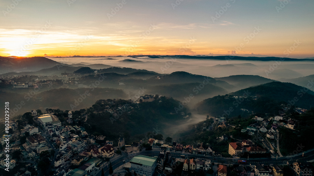 Fototapeta premium Aerial view of Dalat City at dawn, showcasing mist-covered mountains, lush greenery, scattered buildings, and serene atmosphere creating a picturesque landscape
