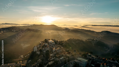 Aerial view of Da Lat at sunrise, showcasing mist-covered buildings amidst lush mountains, under a golden sky, highlighting the city’s architectural and natural beauty.