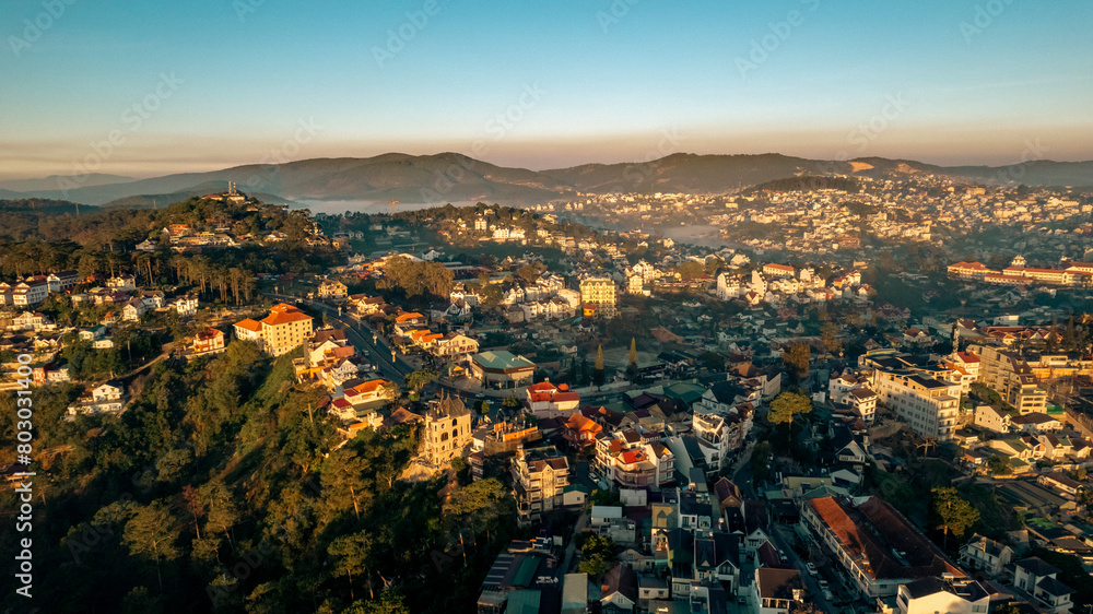 Aerial view of Dalat City at sunrise, showcasing urban landscape amidst natural beauty, buildings nestled between lush green hills and misty valleys, under the golden hour light