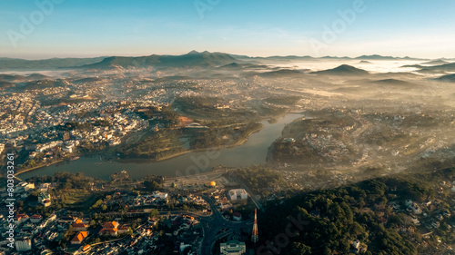 Aerial view of Dalat City at sunrise, featuring the tranquil Xuan Huong Lake amidst urban structures, surrounded by lush greenery and misty hills under a golden sky.