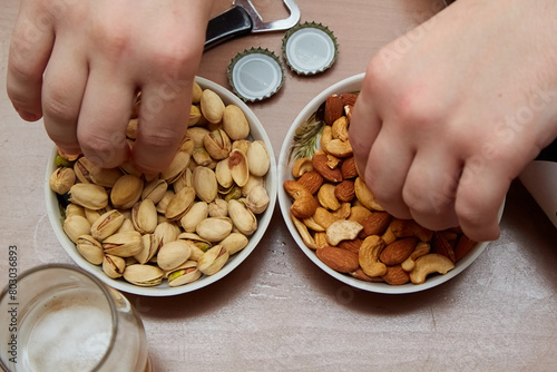 Two hands taking nuts from plates next to a beer glass and a bottle opener, beer snacks, nut snacks, salted pistachios and cashews for beer