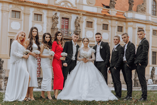 A group of people are posing for a picture in front of a building. The bride and groom are in the center of the group, with the bride wearing a white dress and the groom wearing a suit