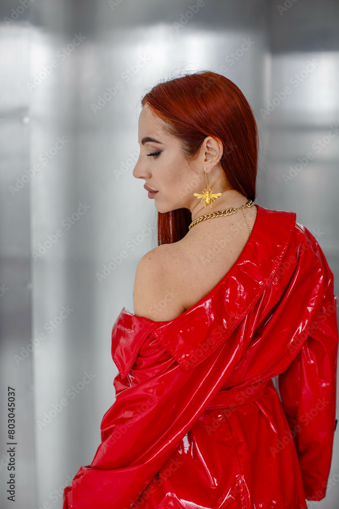 A beautiful young red-haired girl, sexy in a red cloak, stands with her shoulder bare in the studio on a silver background. Back view