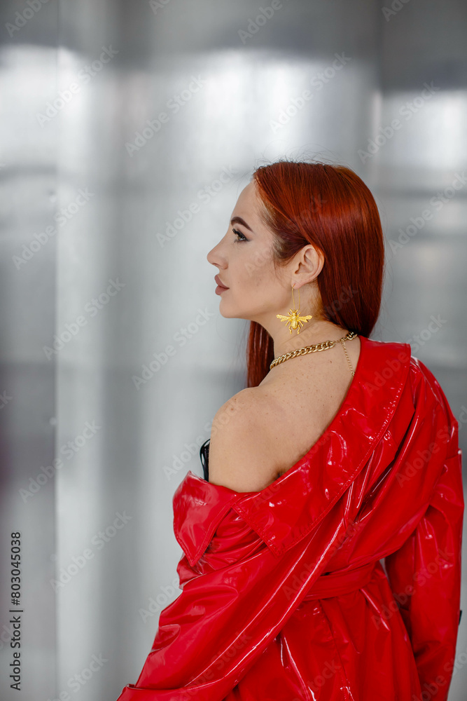 A beautiful young red-haired girl, sexy in a red cloak, stands with her shoulder bare in the studio on a silver background. Back view
