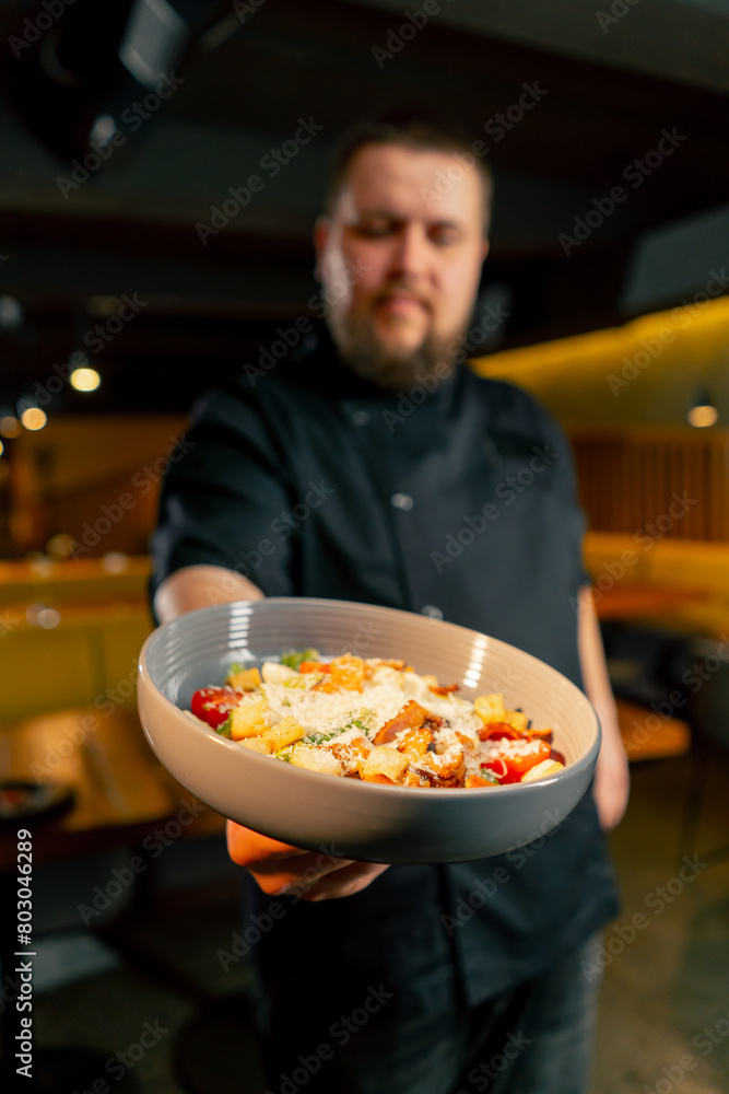 close up in a restaurant a chef in a black jacket stands smiling with a prepared salad