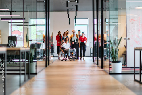 A diverse group of young business people walking a corridor in the glass-enclosed office of a modern startup, including a person in a wheelchair and a woman wearing a hijab