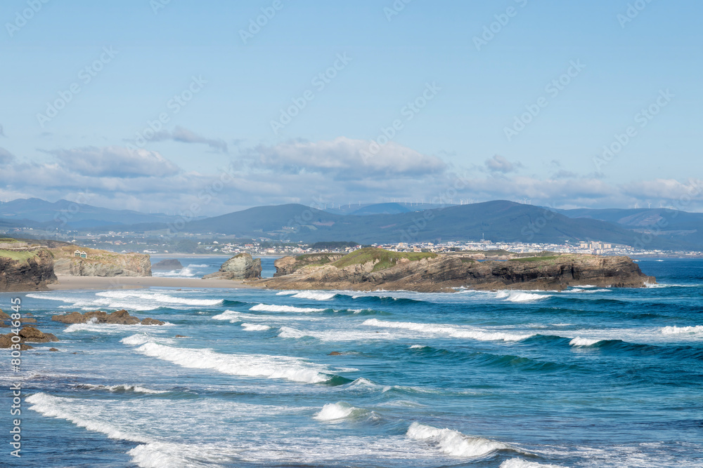 a scenic coastline with waves crashing against rocky cliffs, framed by mountains and a clear sky