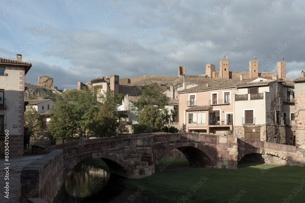 a historic town with stone buildings, an arched bridge, and distant castles under a partly cloudy sky
