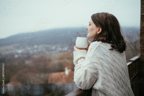 Young woman in white knitted sweater holding white cup of tea, standing on a balcony of a wooden house with mountains view.