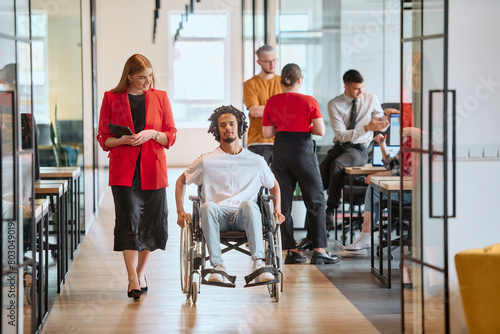 A business leader with her colleague, an African-American businessman who is a disabled person, pass by their colleagues who work in modern offices