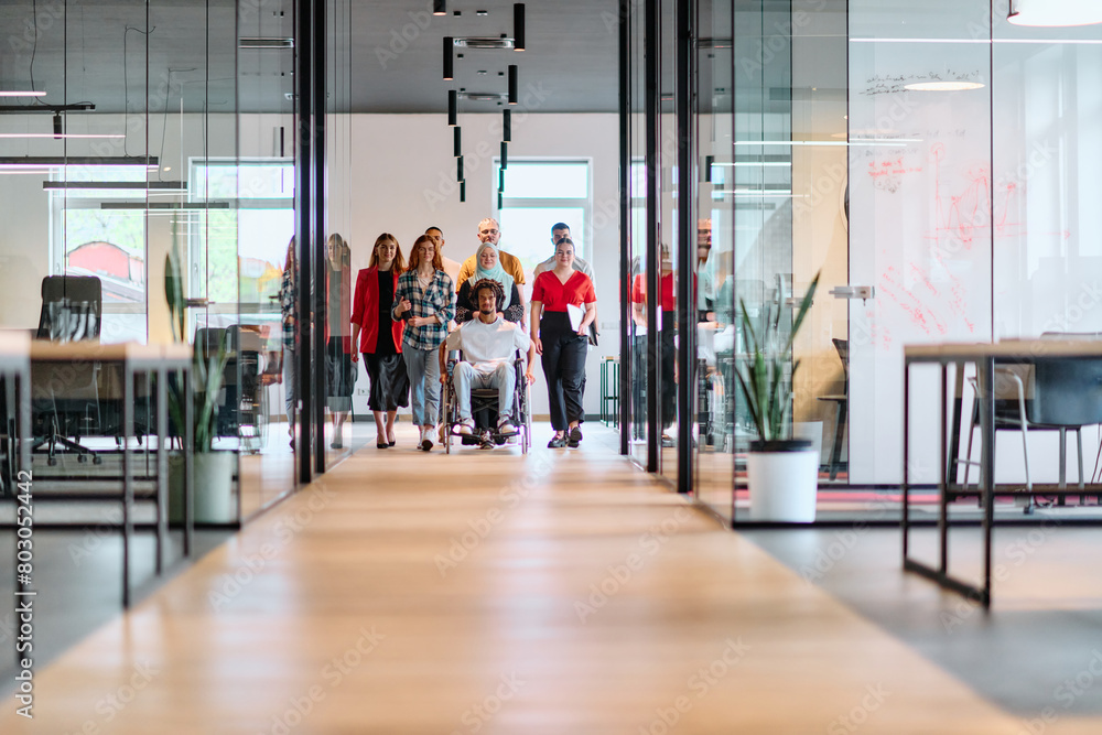 A diverse group of young business people walking a corridor in the glass-enclosed office of a modern startup, including a person in a wheelchair and a woman wearing a hijab