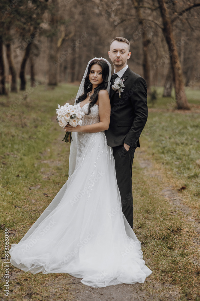 A bride and groom are posing for a picture in a forest. The bride is wearing a white dress and holding a bouquet of flowers. Scene is romantic and intimate