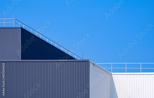 Gray and white aluminium corrugated factory building in geometric minimal style with steel fence on rooftop against blue clear sky background