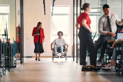 A business leader with her colleague, an African-American businessman who is a disabled person, pass by their colleagues who work in modern offices