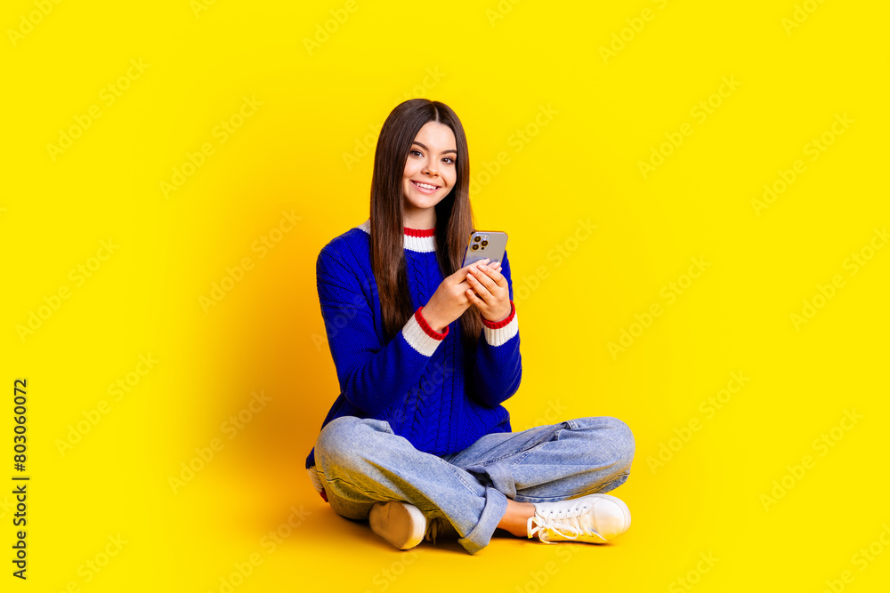 Full length photo of pretty teen girl hold device crossed legs wear trendy knitwear blue outfit isolated on yellow color background Full length photo of pretty teen girl hold device crossed legs wear trendy knitwear blue outfit isolated on yellow color background