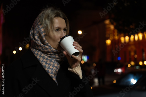 Young beautiful woman in a stole and a black coat drinks takeaway coffee from a paper cup on a lighted city street at night. photo