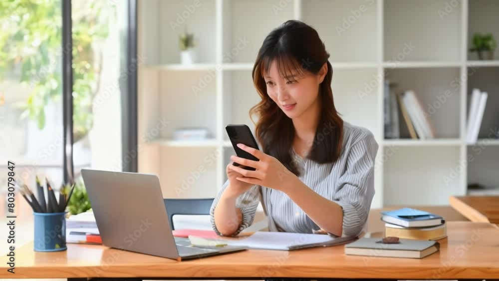 Smiling young Asian woman sits at wooden desk, focusing on smartphone screen while studying or working remotely on laptop computer at home. Smiling young Asian woman sits at wooden desk, focusing on smartphone screen while studying or working remotely on laptop computer at home.