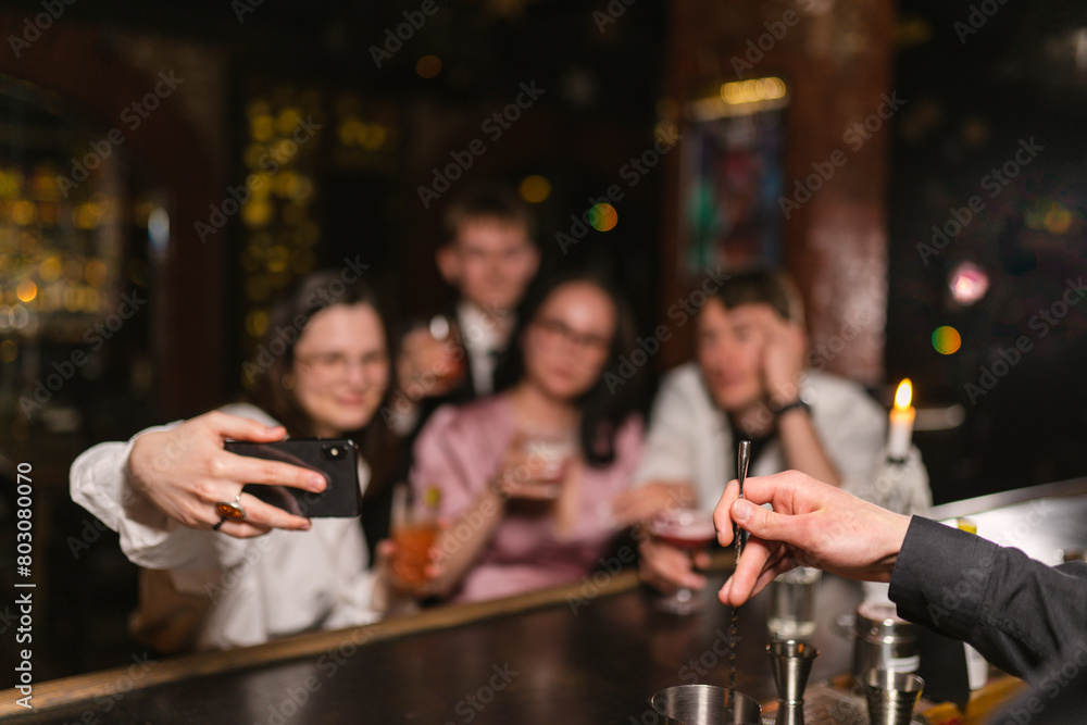 Happy guests group makes selfie and bartender makes beverage at counter focus on hands. Fancy nightlife scene in cosy pub