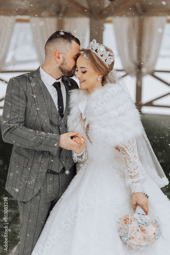 A bride and groom are standing in the snow, with the bride wearing a white fur coat. They are holding hands and smiling at the camera