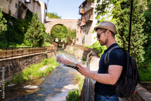 A young man explores new destinations by traveling through North Macedonia.He is standing by the river and looking at the map.