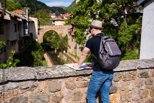 A young man explores new destinations by traveling through North Macedonia.He is standing on the bridge and looking at the map.