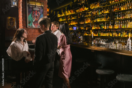 Young guys and ladies speak and drink alcohol in modern pub. Atmospheric pub with variety of alcoholic bottles and subdued lamps