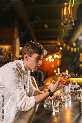 Guy in white shirt sits among empty glasses at bar counter in cozy pub. Guy holds bottle with expensive alcohol in hand and wants drink