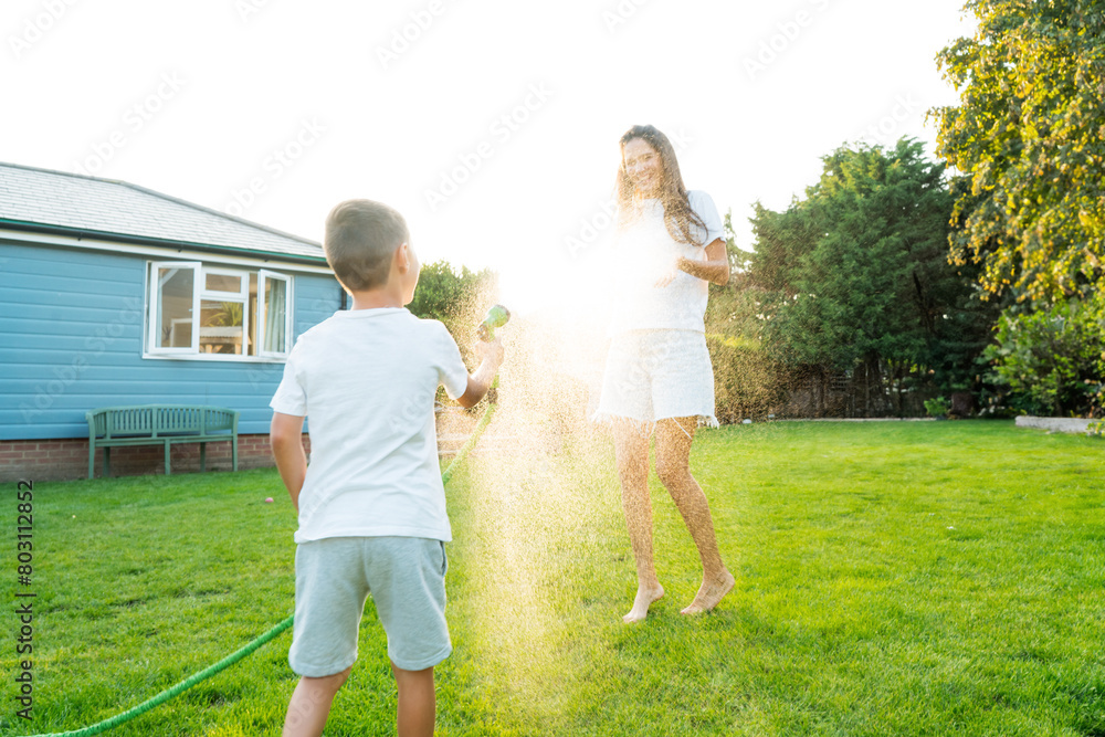 Joyful kid with mother playing with water sprinkler hose, splashing, laughing, having fun together in garden on sunset. Summer outdoor activity. Happy active childhood, family time. Selective focus