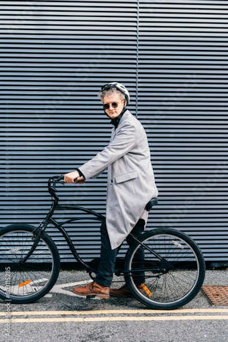 Stylish male in coat, sunglasses and protective helmet riding on retro bicycle near gray urban wall. Neutral carbon footprint transportation. Green eco friendly mobility sustainable transport.