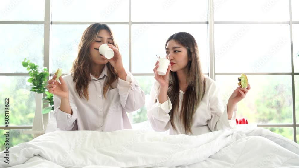 Two beautiful Asian girls are roommates wearing a white shirt, sitting on a bed holding a coffee mug in a bedroom in an apartment. Two beautiful Asian girls are roommates wearing a white shirt, sitting on a bed holding a coffee mug in a bedroom in an apartment.