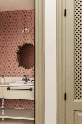 Bathroom with cream marble countertop, gold-framed mirrors, pink wallpaper and white porcelain sink and a built-in wardrobe with lattice doors in an antechamber