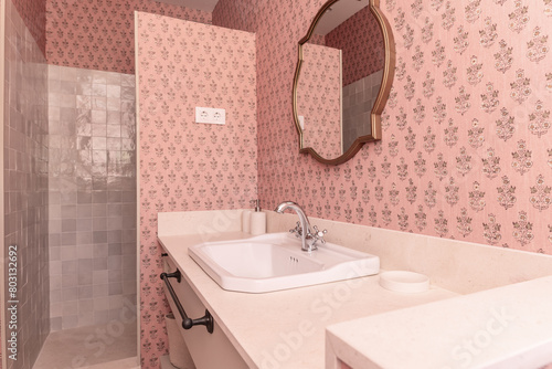 Bathroom with cream marble countertop, gold-framed mirrors, pink wallpaper, a light-tiled walk-in shower, and white porcelain sink